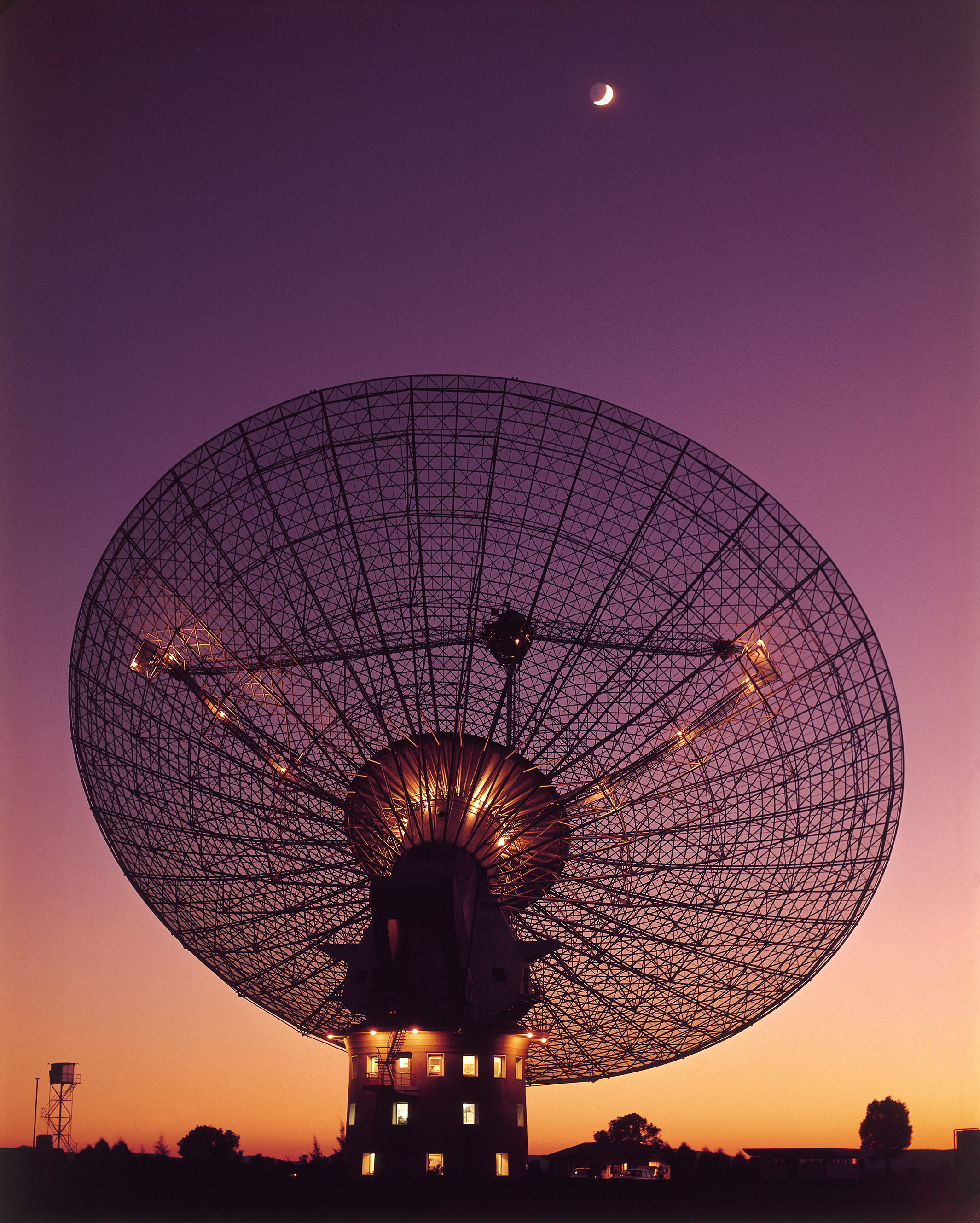 The silhouette of a large radio telescope dish at sunset. The Moon is high in the sky above it.